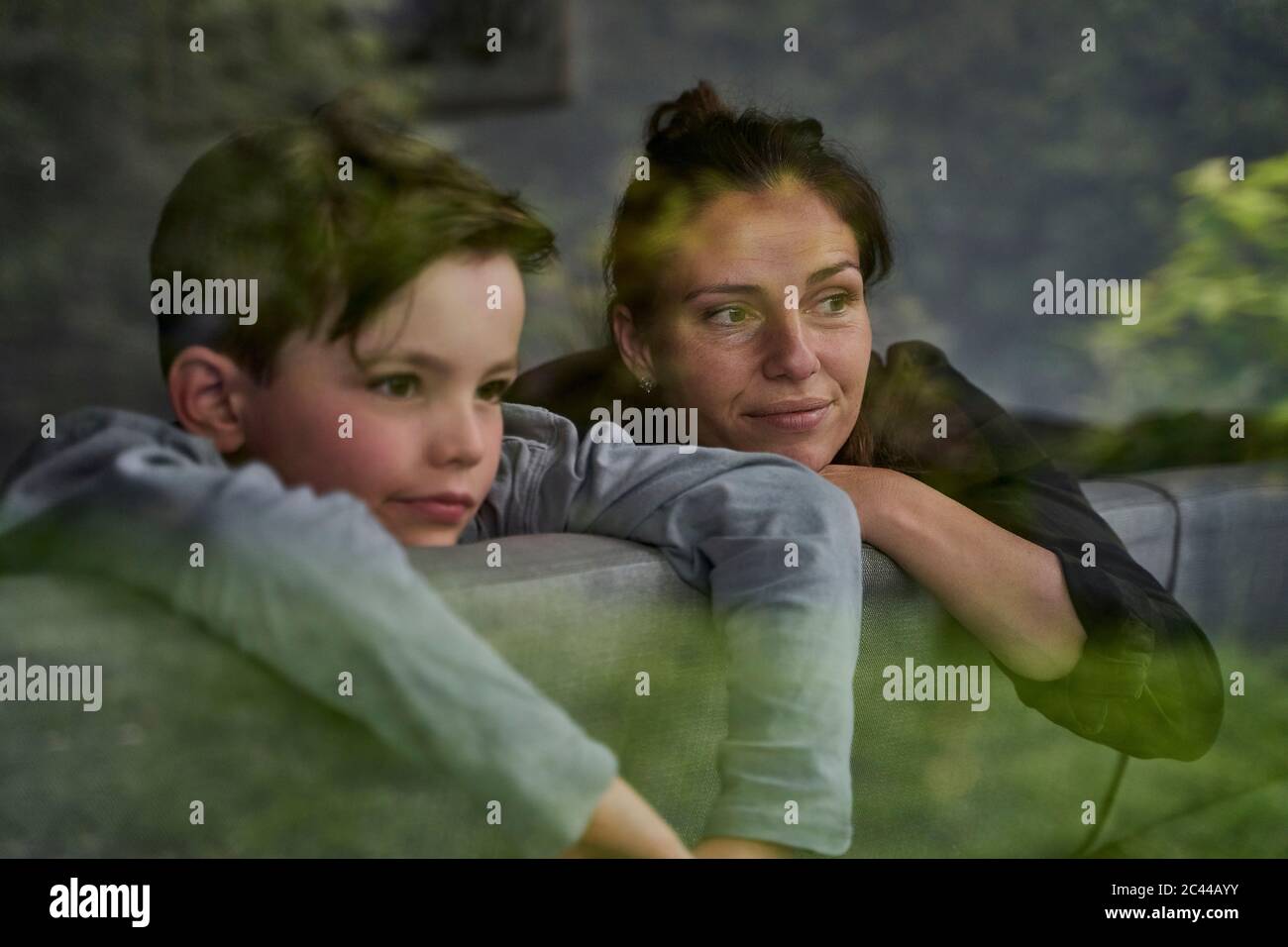 Nachdenkliche Mutter und Sohn blicken weg, während sie sich auf dem Sofa zu Hause entspannen, durch das Fenster gesehen Stockfoto