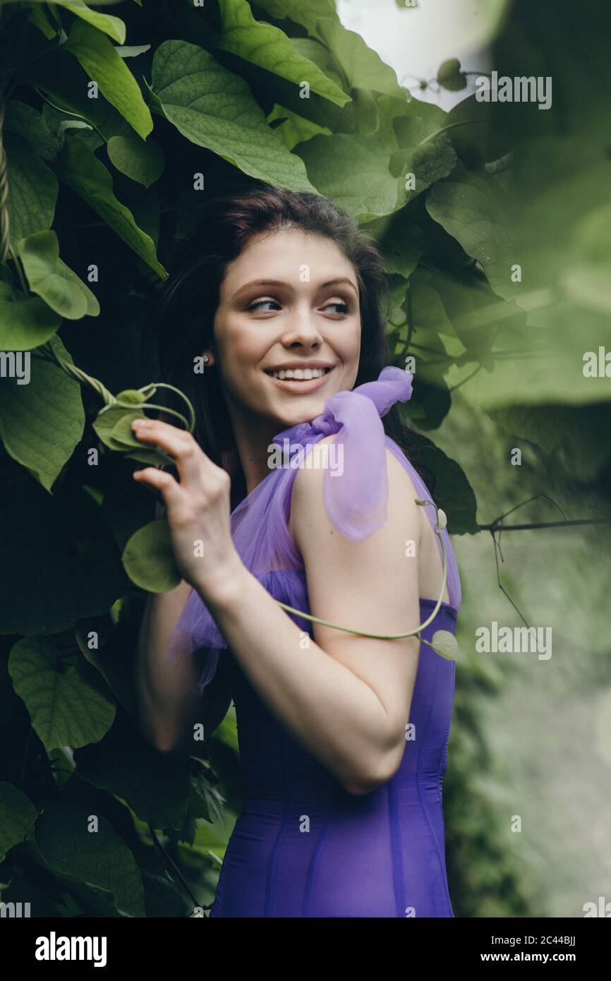 Junge lächelnde Frau in violettem Kleid, die in der Nähe der grünen Wand steht Stockfoto