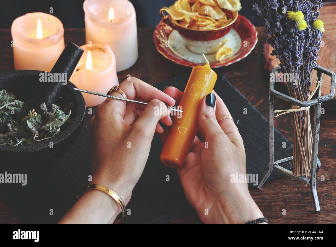 Wiccan Hexe schnitzt Symbole und Sigillen auf gelbgoldfarbenen Kerzenständer an ihrem Altar. Kerze in den Händen halten. Hexenelemente in unscharfem Hintergrund Stockfoto