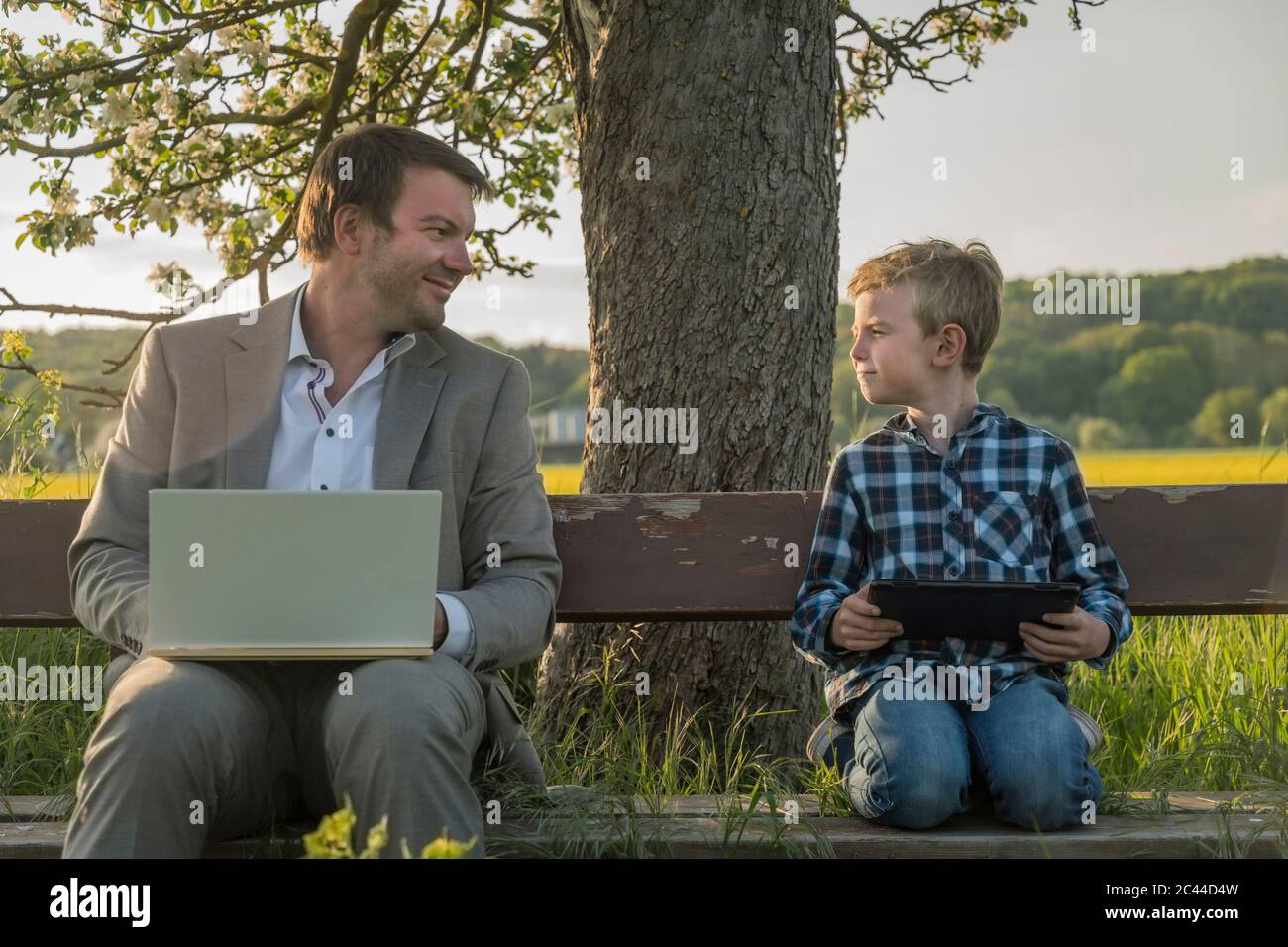 Lächelnder Mann, der den Sohn mit Laptop und digitalem Tablet auf der Bank ansieht Stockfoto