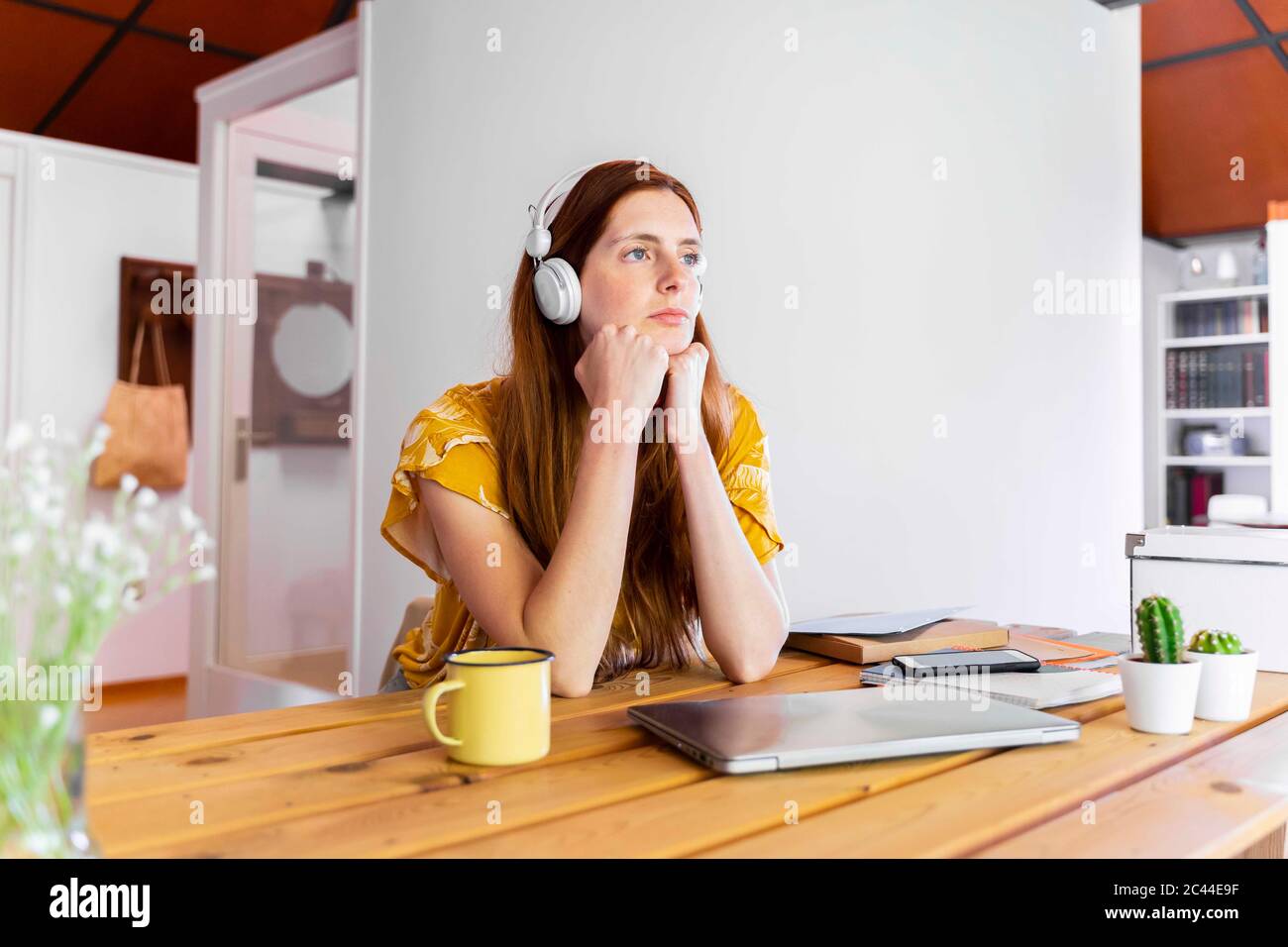 Nachdenkliche junge Frau mit Händen am Kinn, die an der Wand am Schreibtisch sitzt Stockfoto