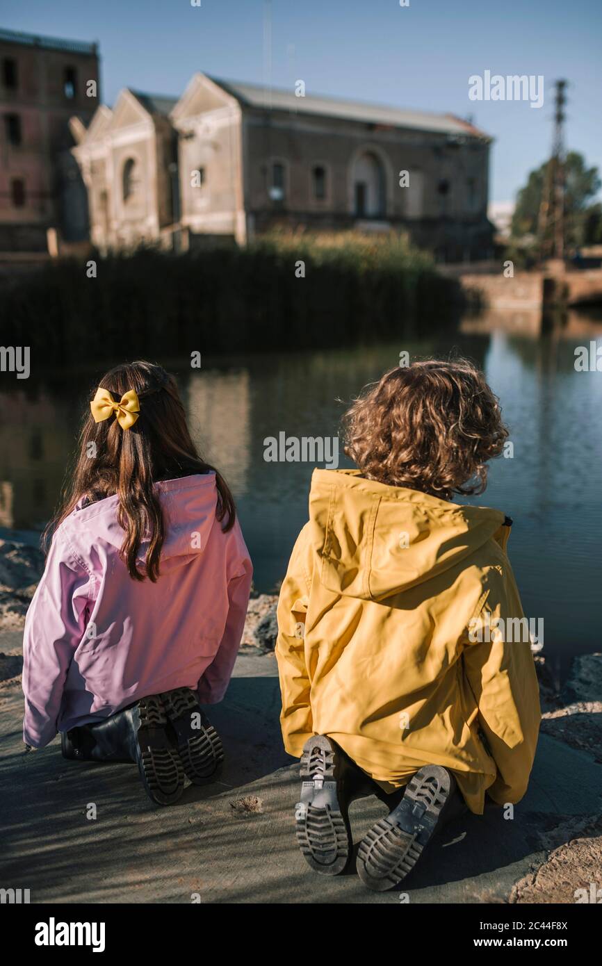 Geschwister tragen Regenmäntel beim Knien am Flussufer Stockfoto