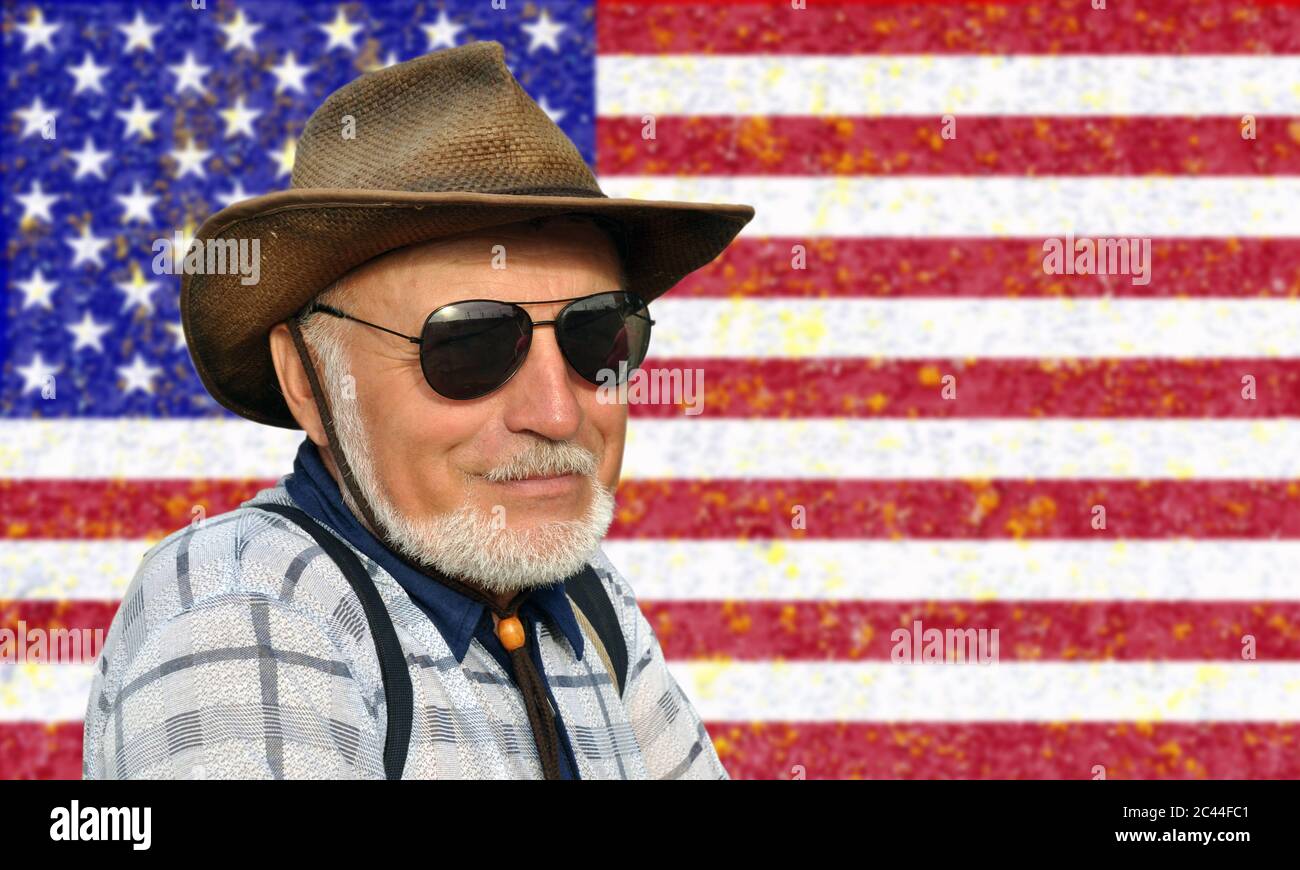Porträt eines nicht jungen Mannes in einem Cowboy-Hut und Brille mit einem grauen Bart vor dem Hintergrund der USA-Flagge. Stockfoto