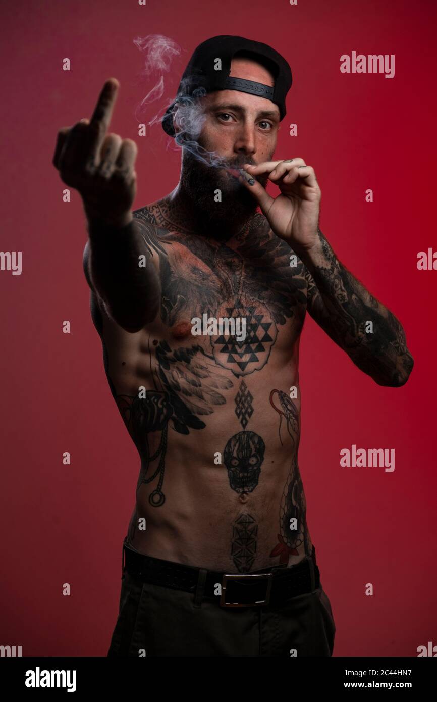 Portrait von tätowierten Mann vor rotem Hintergrund geben den Finger beim Rauchen ein Gelenk Stockfoto