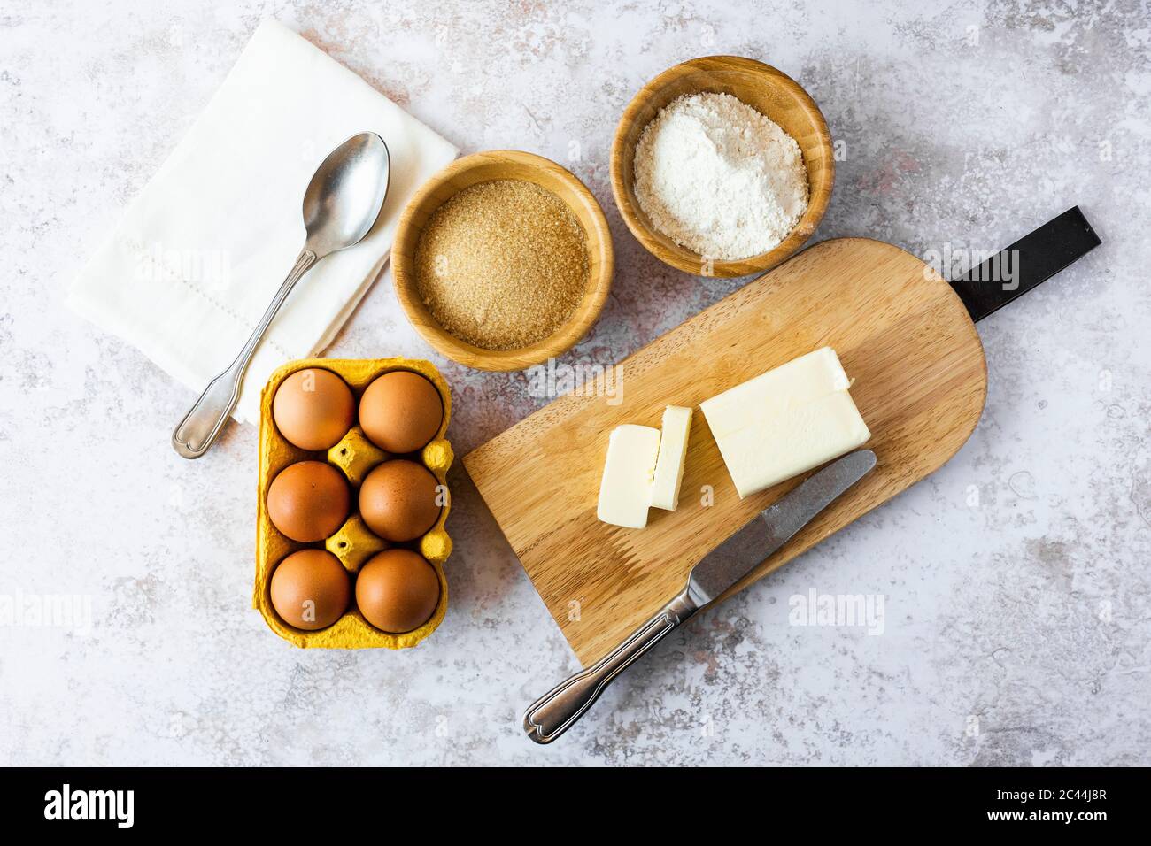 Serviette, Löffel, Schalen mit Mehl und braunem Zucker, Hühnereier, Schneidebrett, Butter und Tafelmesser Stockfoto