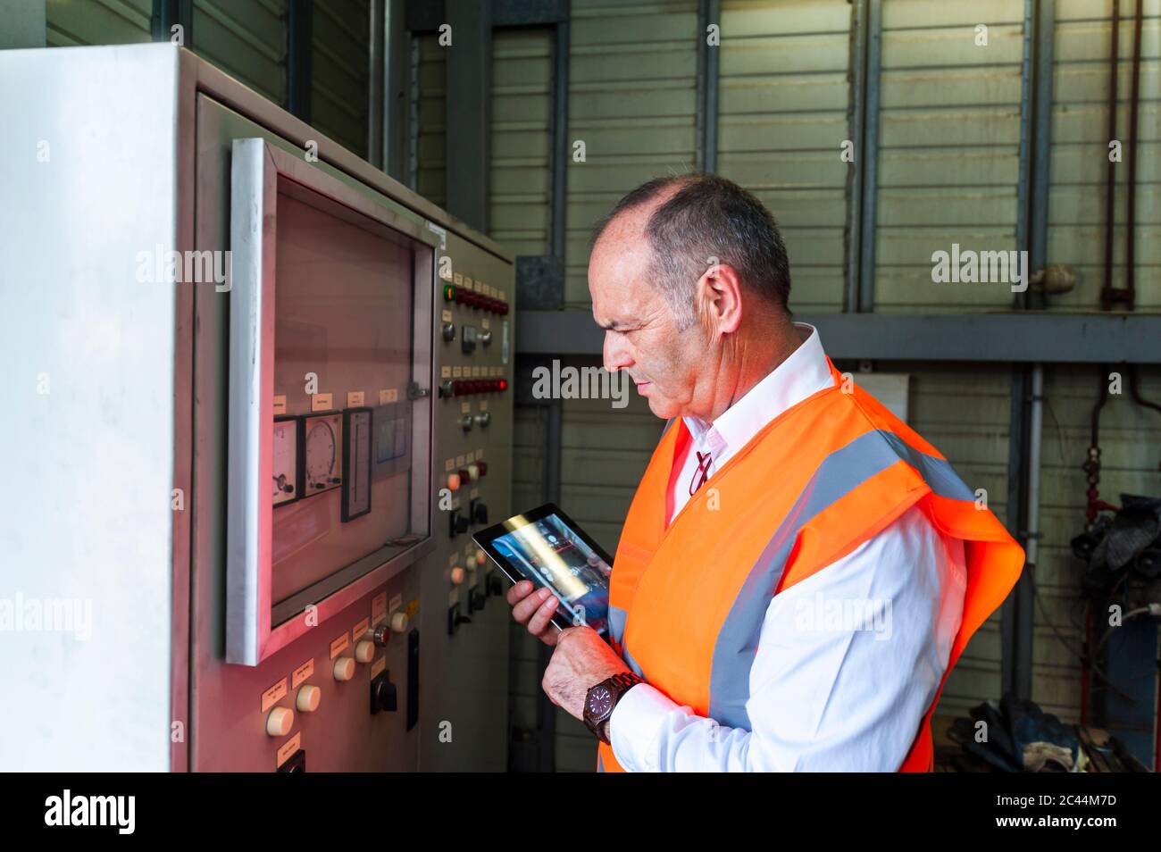 Älterer Mann mit Tablet, der eine Sicherheitsweste trägt und eine Maschine untersucht Stockfoto