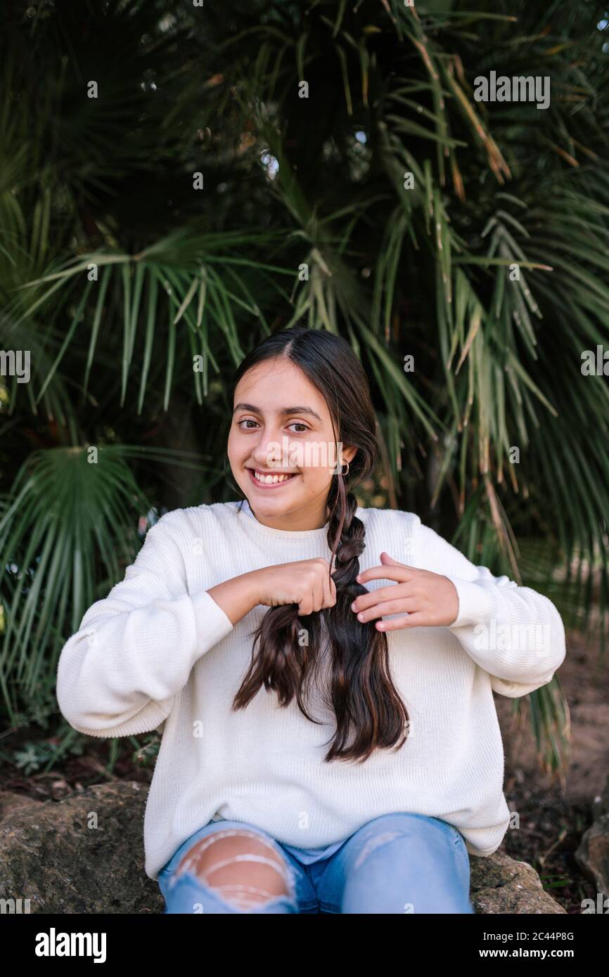 Portrait eines lächelnden Teenagers, das im Park gegen die Palme sitzt und ihr langes braunes Haar flechtet Stockfoto