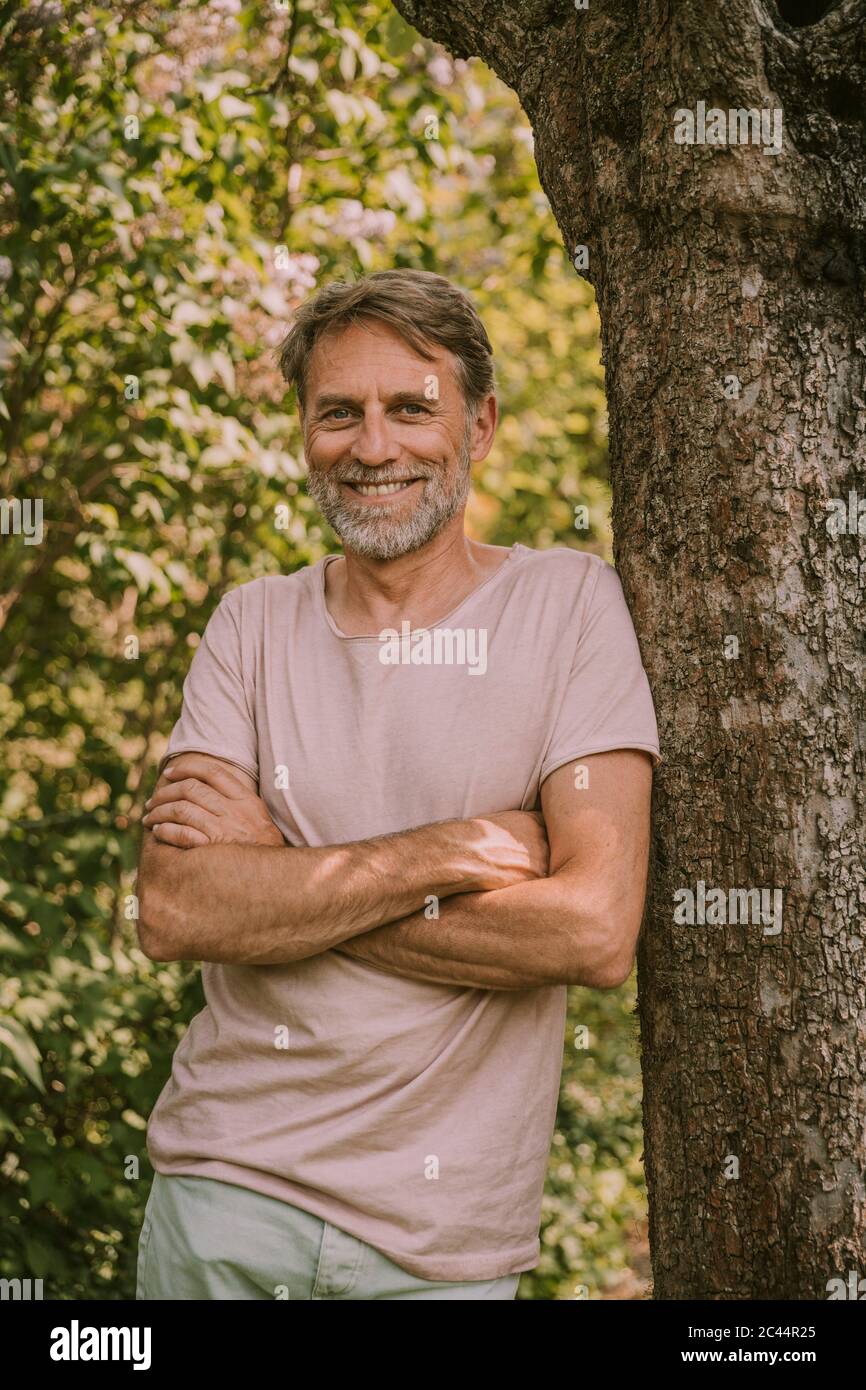 Lächelnder bärtiger reifer Mann, der mit gekreuzten Armen auf Baumstamm im Garten lehnt steht Stockfoto