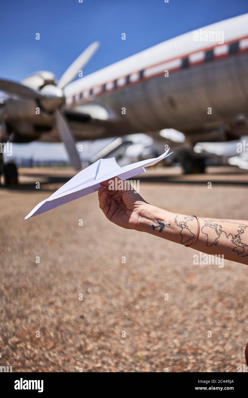 Die Hand der Frau mit Tätowierung, die Papierflugzeug gegen Luftfahrzeug am Flughafen hält, an sonnigen Tagen Stockfoto