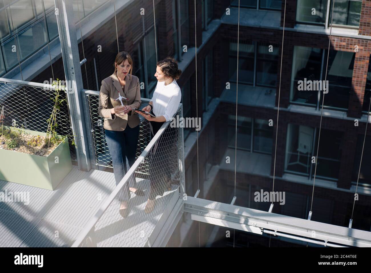 Zwei Geschäftsfrauen mit Tablet und Windturbinenmodell, die auf dem Balkon eines Bürogebäudes eine Besprechung abhalten Stockfoto