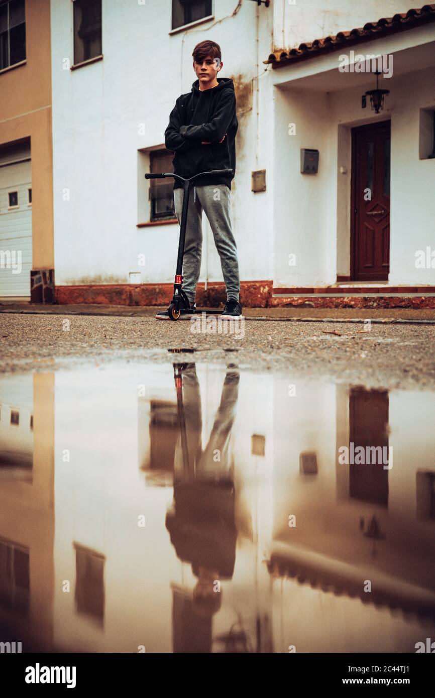 Teenager Junge steht mit Push-Scooter auf der Straße mit Pfütze im Vordergrund Stockfoto