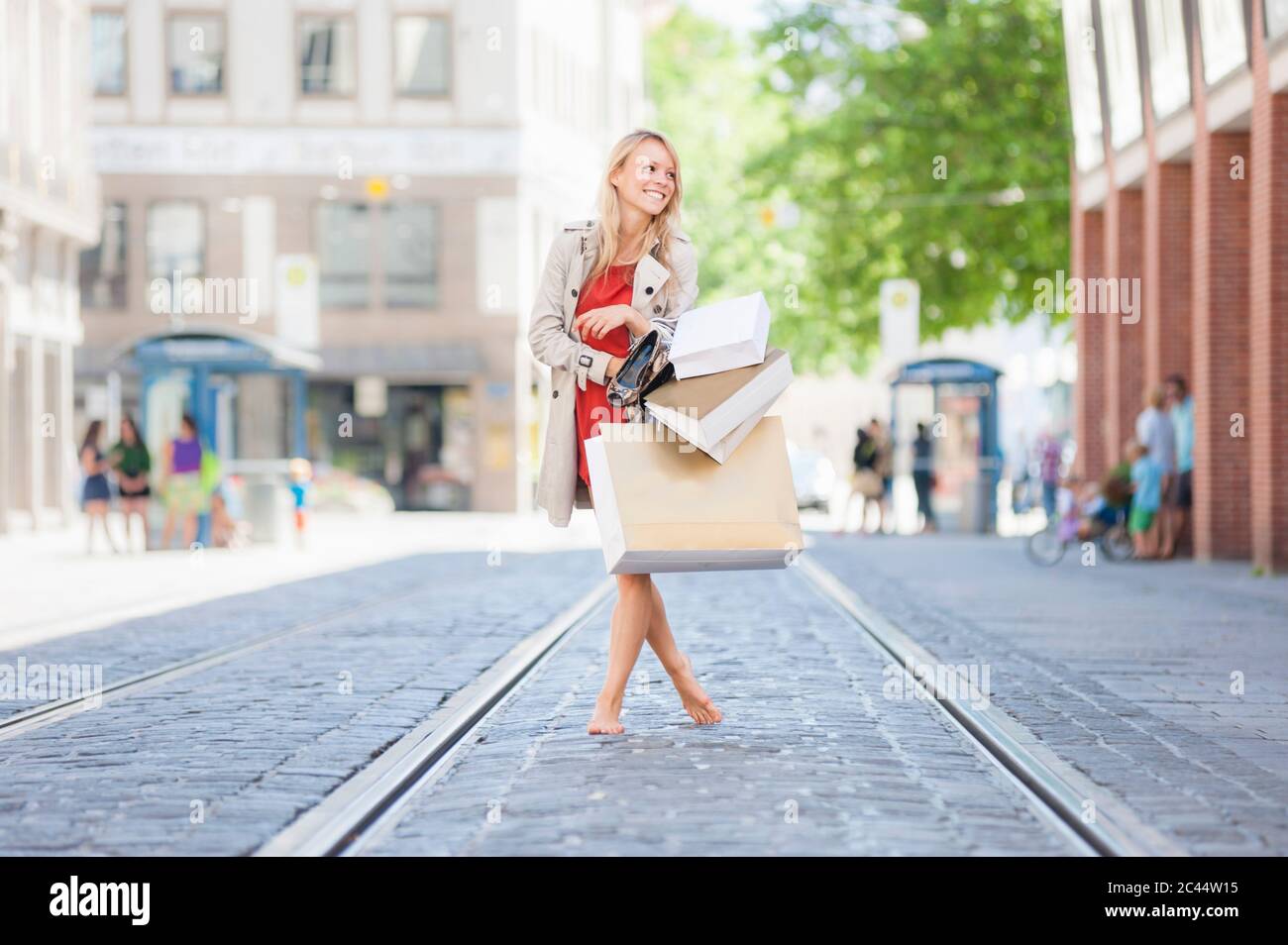 Lächelnde Frau, die Schuhe und Einkaufstaschen hält, während sie barfuß auf der Straße läuft Stockfoto