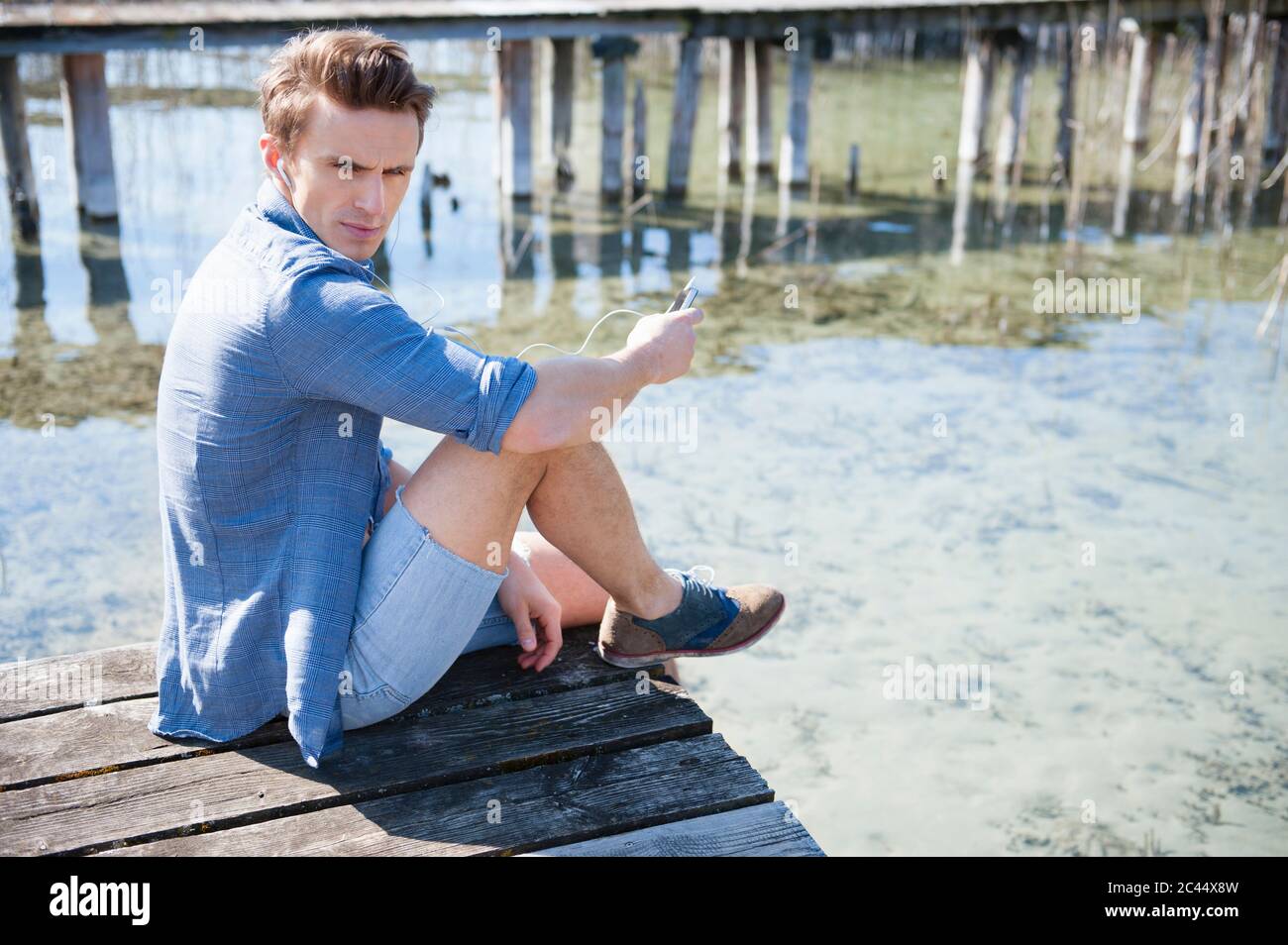 Schöner Mann mit mittlerem Erwachsenen, der Smartphone benutzt, während er auf dem Pier über dem See sitzt Stockfoto