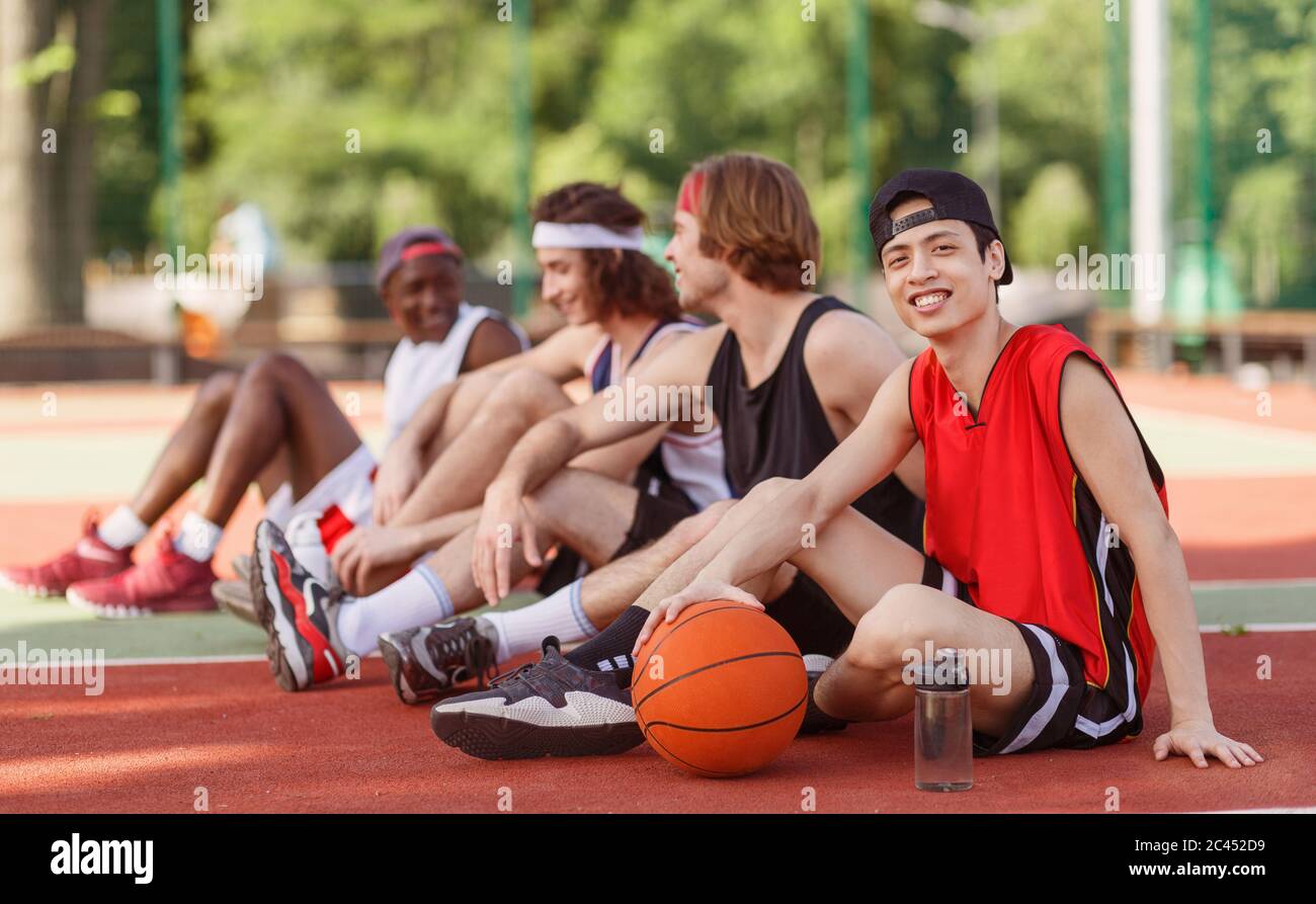 Lächelnder asiatischer Basketballspieler mit seinem vielfältigen Team, das auf dem Freiluftspielplatz sitzt, Platz zum Kopieren Stockfoto