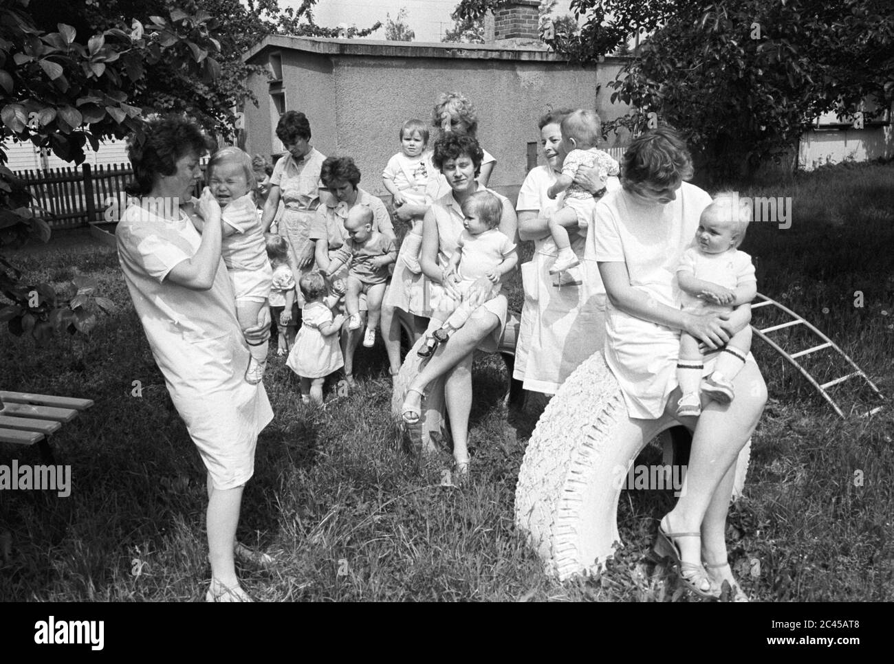 12. August 1984, Sachsen, Rackwitz: Im Garten des crèche in Rackwitz beschäftigen sich die crèche-Pädagogen Mitte der 1980er Jahre mit den Kindern. Sie sitzen auf alten LKW-Reifen, die weiß lackiert und in den Boden eingebettet und zum Klettern verwendet wurden. Ihre Eltern arbeiten hauptsächlich im VEB Leichtmetallwerk Rackwitz und in der Landwirtschaft. Genaues Aufnahmedatum nicht bekannt. Foto: Volkmar Heinz/dpa-Zentralbild/ZB Stockfoto