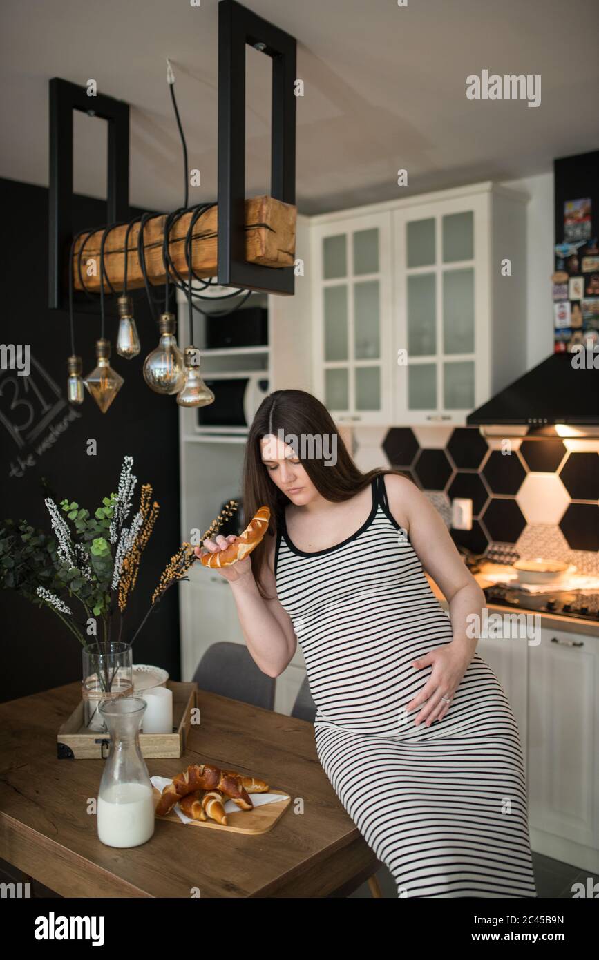 Junge schwangere Frau in ihrer Küche essen einen Muffin. Schwangere Frau in der Küche sitzt an einem Tisch und isst Stockfoto