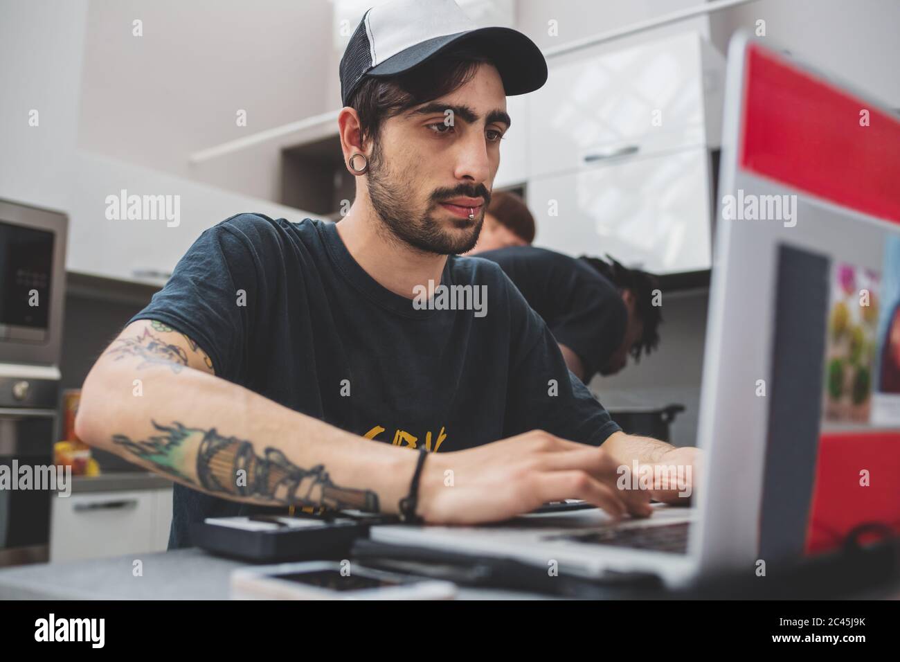 Junger bärtiger Mann mit Piercings, der an einem Tisch sitzt, Baseballmütze trägt und auf dem Laptop tippt. Stockfoto