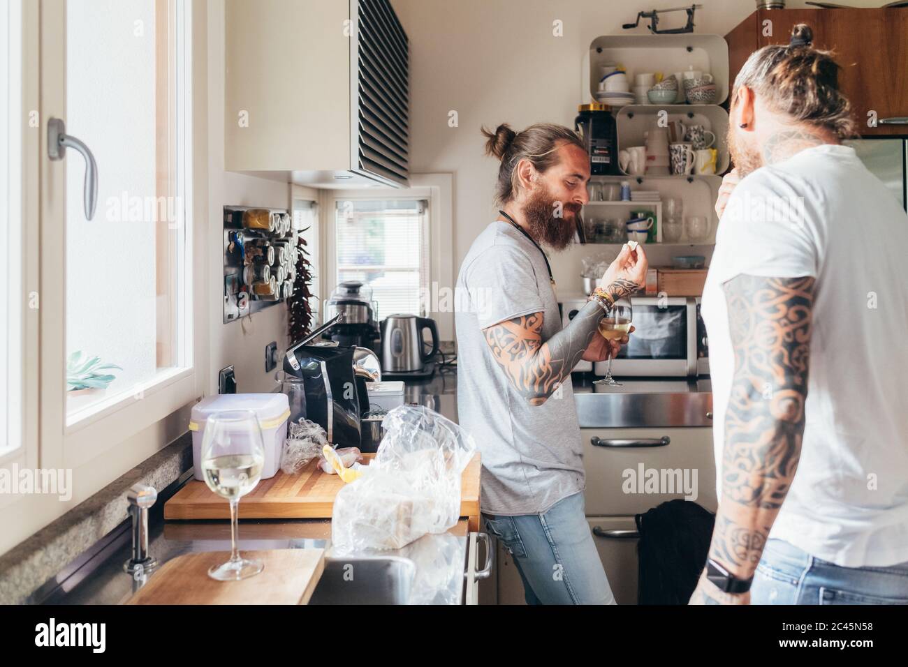 Zwei bärtige tätowierte Männer mit langen Brünetten Haaren stehen in einer Küche und essen. Stockfoto
