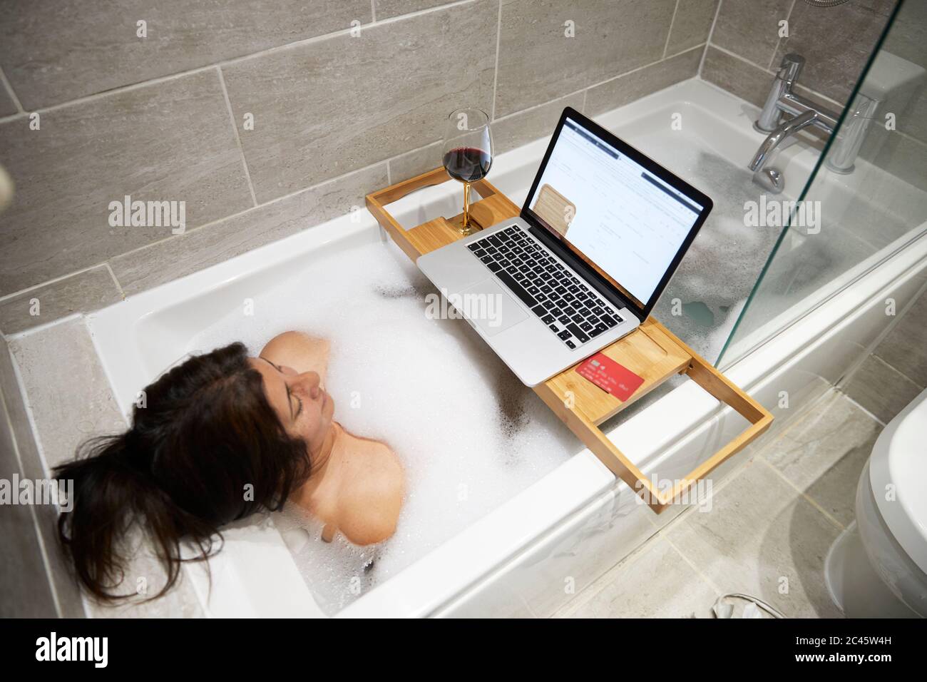 Frau, die in der Badewanne sitzt, ein Schaumbad hat und während der Coronavirus-Krise am Laptop arbeitet. Stockfoto