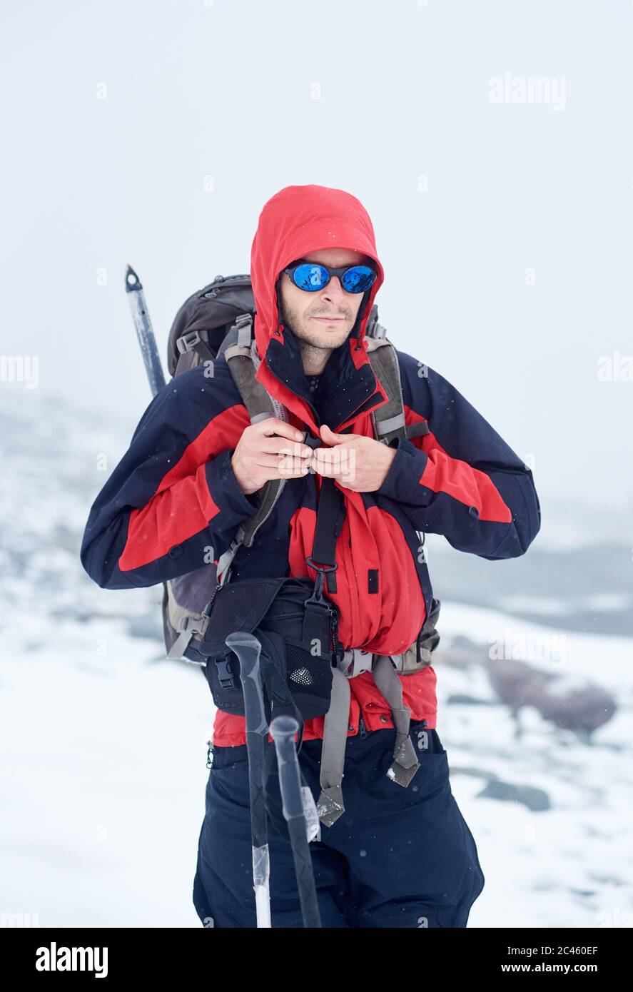 Männliche Tourist in Sonnenbrille Befestigung Reise Rucksack und Blick auf Kamera. Wanderer mit Umhängetasche trägt Sport-Winterjacke mit Kapuze und modischen Farbtönen. Konzept von Reisen, Wandern und Alpinismus Stockfoto