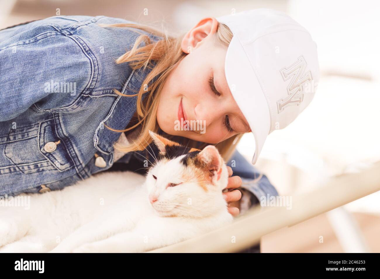 Junge blonde Mädchen, Grundschulalter, trägt Sonnenkappe, streicheln Katze auf der Sonnenliege liegen Stockfoto