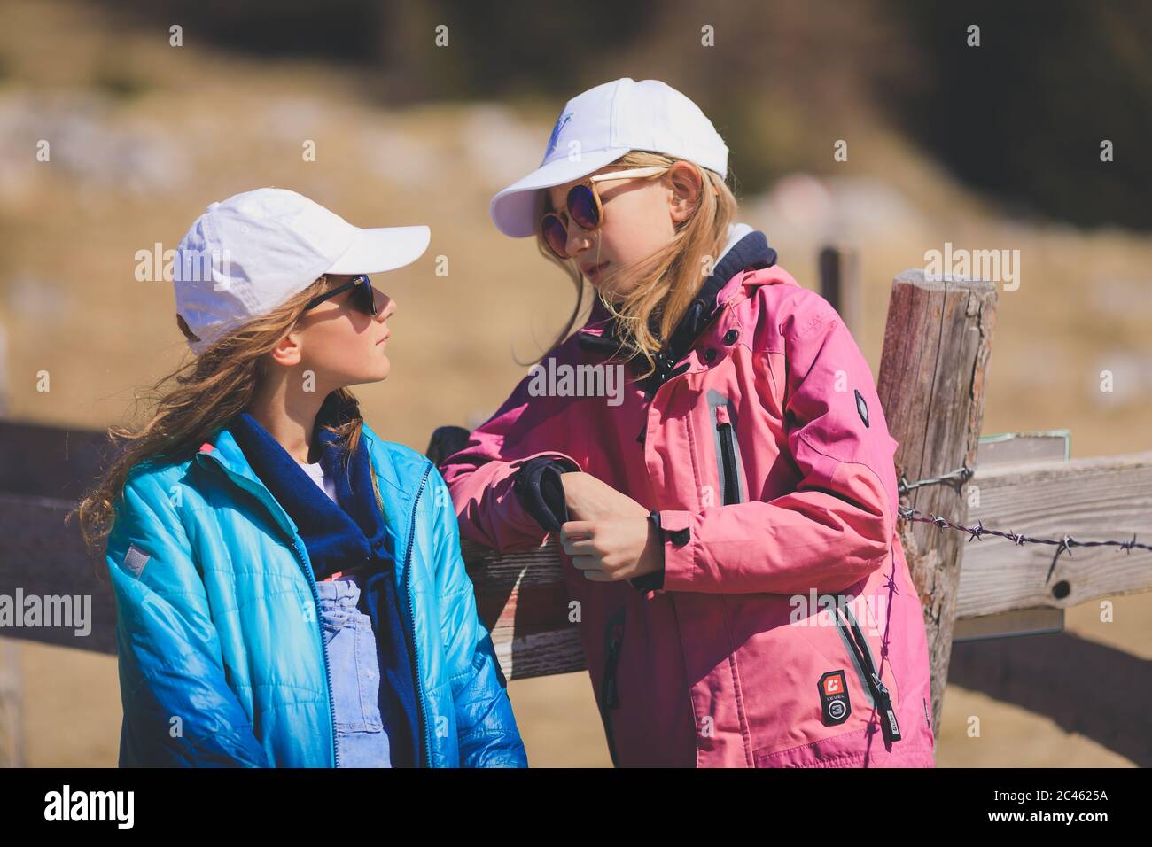 Zwei junge Mädchen, die Sonnenkappen abhauen und sich vor einem Holzzaun unterhalten Stockfoto