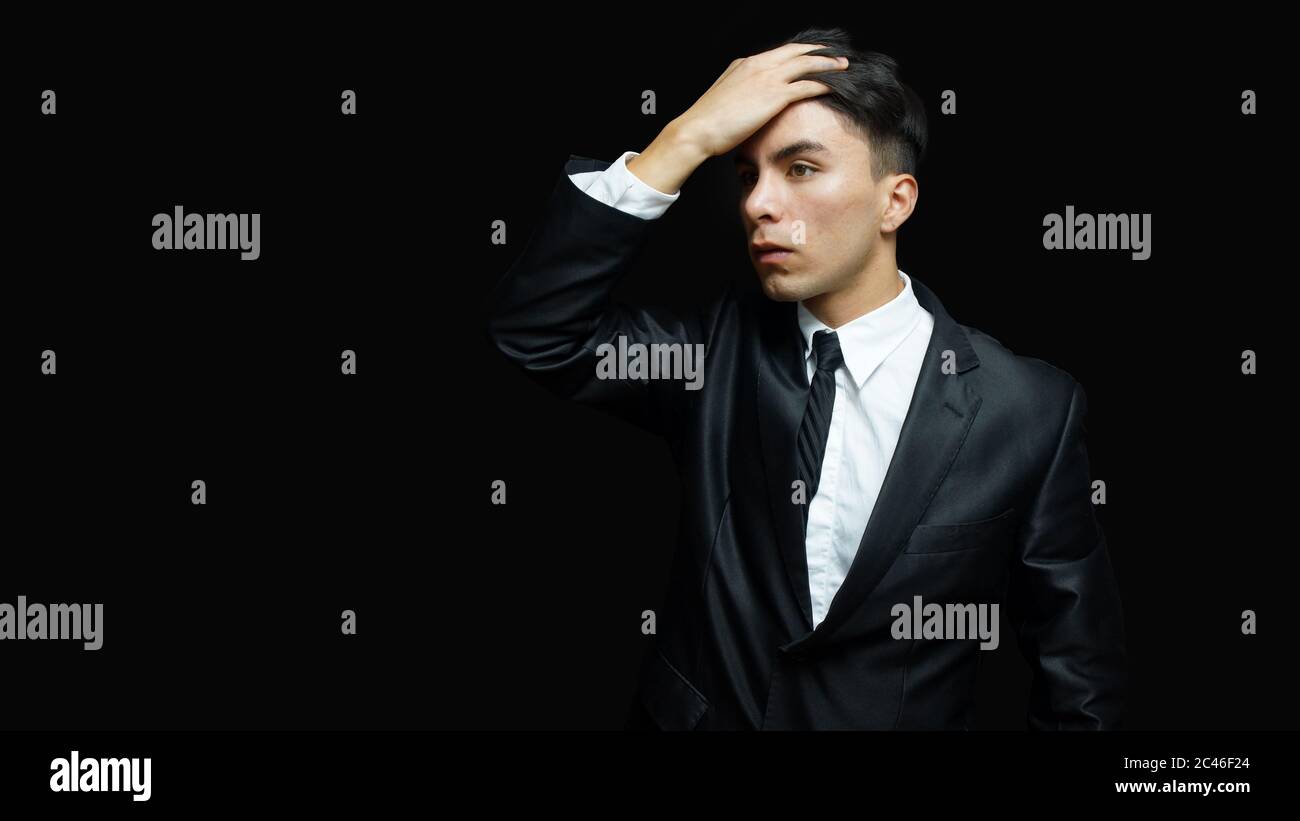 Junger latein in schwarzem Anzug, weißes Hemd, schwarze Krawatte in der Haltung der Sorge mit der Hand auf der Stirn auf schwarzem Hintergrund Stockfoto