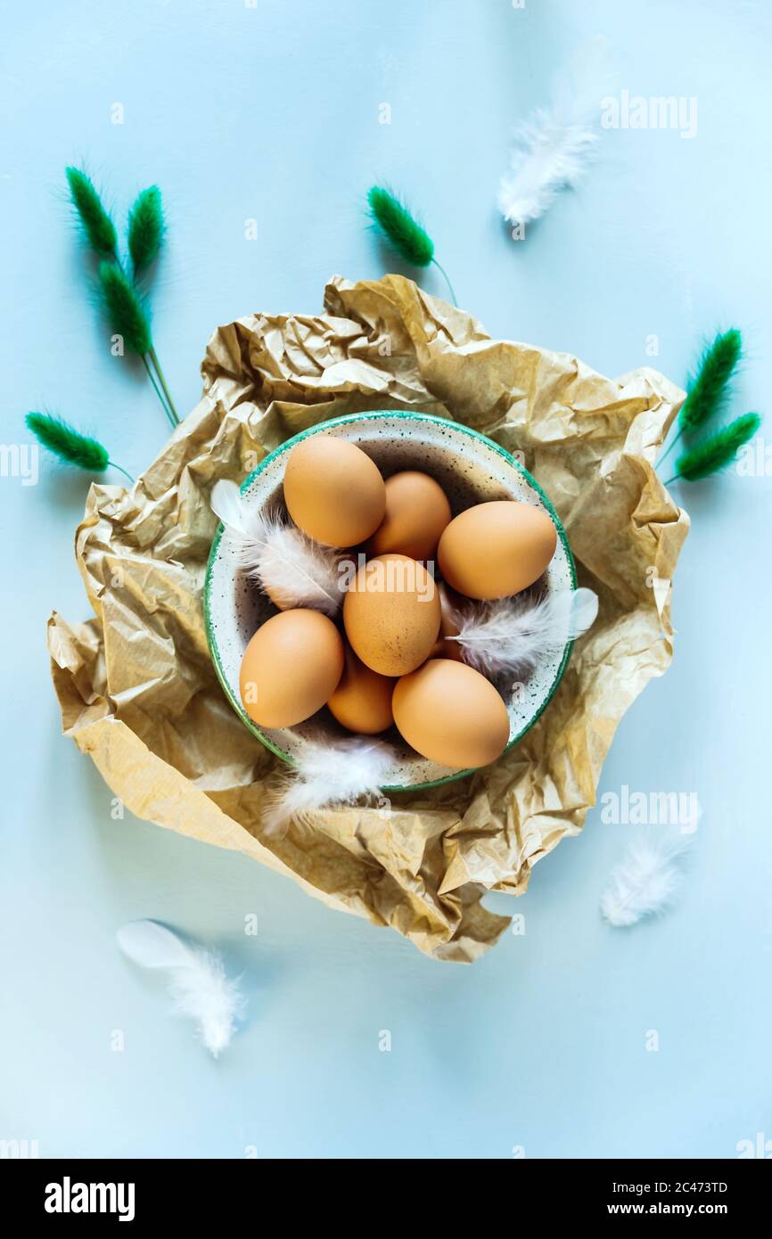 Ein flaches Foto des Osterstilllebens von einer Schale aus braunen Eiern auf zerknittertem Bastelpapier, weißen Federn und trockenen grünen Pflanzen als Dekor. Hellblaue Rückseite Stockfoto