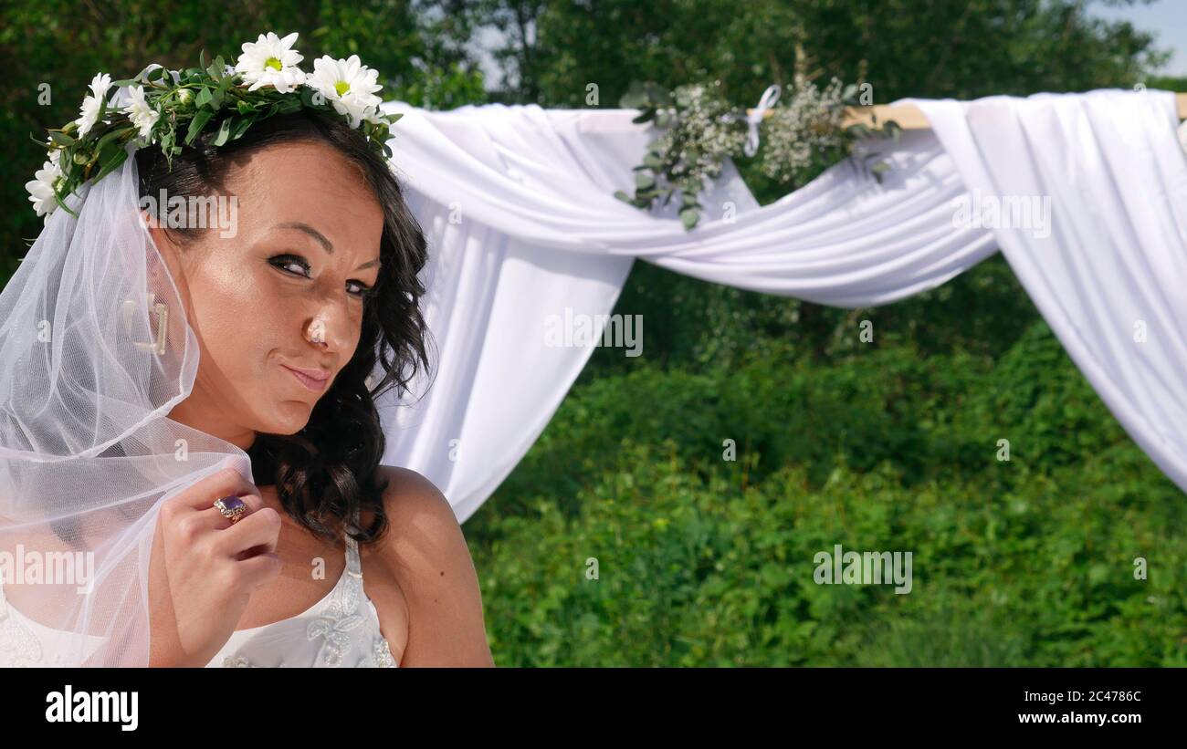 Eine lächelnde Braut mit einem Blumenkranz auf dem Kopf und Schleier vor  dem Hochzeitsbogen Stockfotografie - Alamy