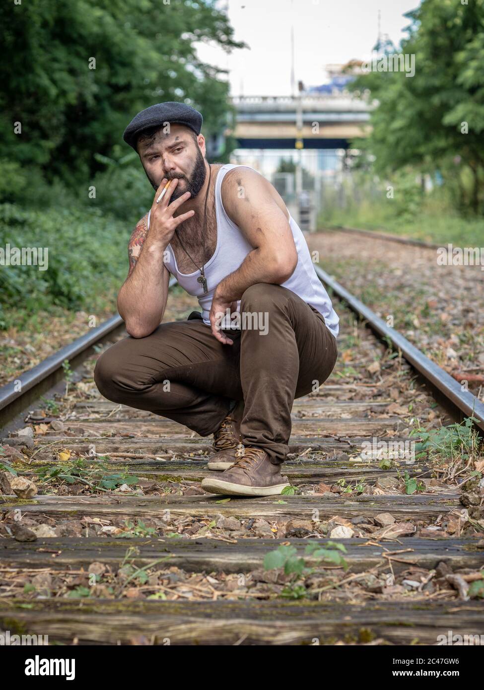 Junger französischer bärtiger Mann, der in Paris, Frankreich, draußen auf einer stillstehenden Eisenbahnstrecke mit natürlichem Licht raucht, mit Tattoos Stockfoto