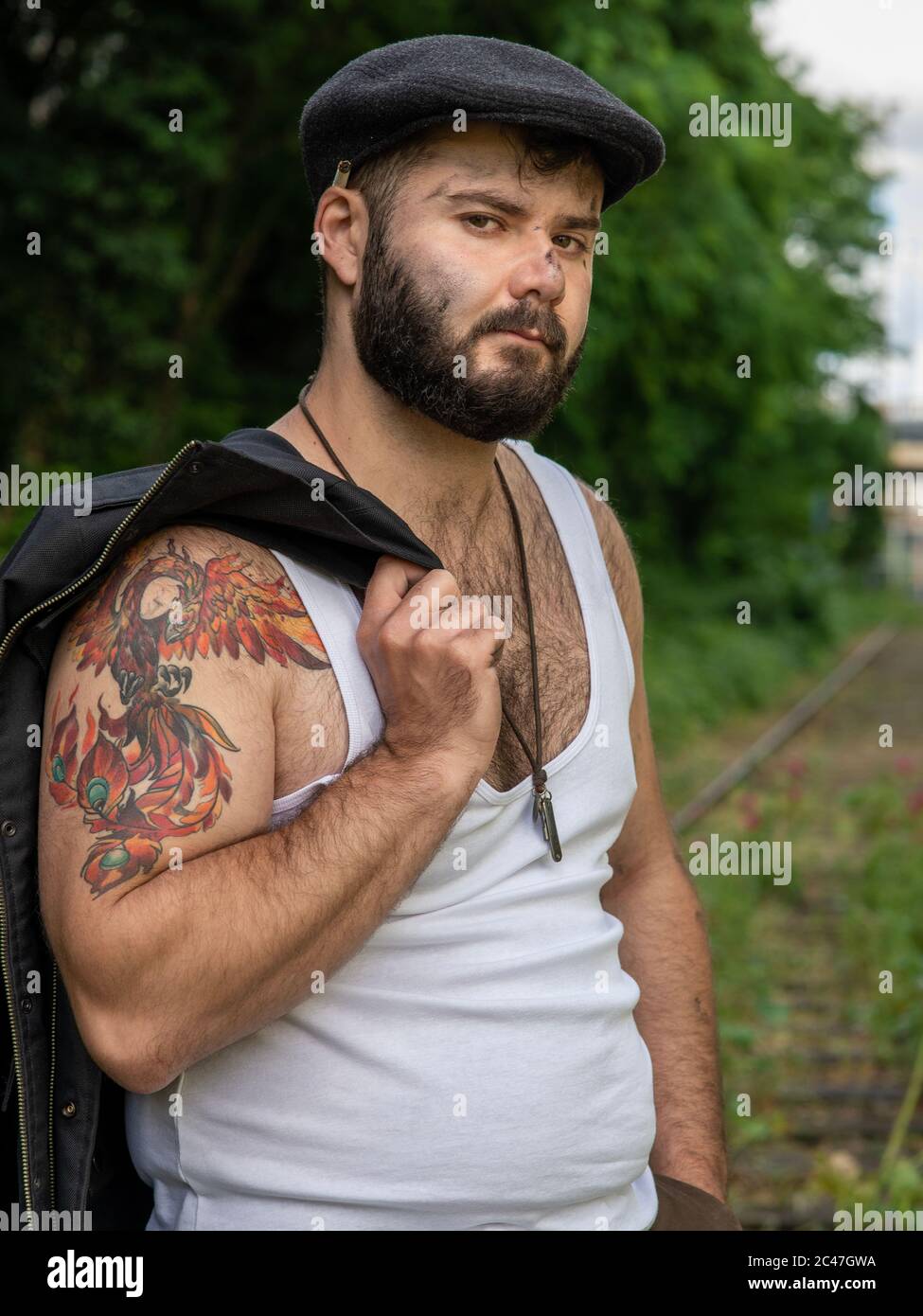 Junger, hübscher, bärtiger Mann mit Tattoos und Kohlestatzen auf der Haut, der auf einer stillstehenden Eisenbahnstrecke mit Blumen mit natürlichen Motiven draußen aufgenommen wurde Stockfoto