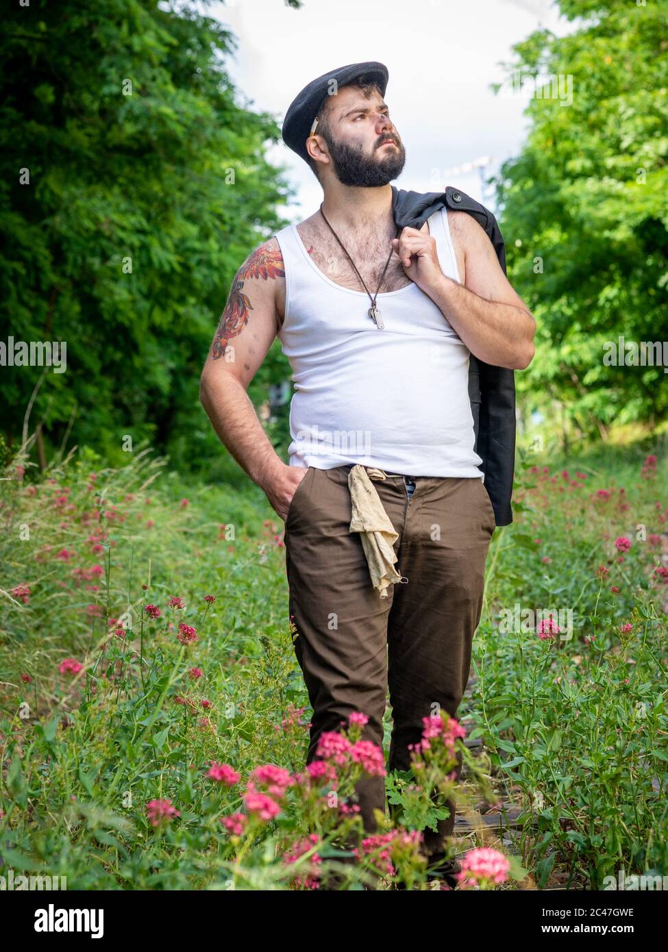 Junger, hübscher, bärtiger Mann mit Tattoos und Kohlestatzen auf der Haut, der auf einer stillstehenden Eisenbahnstrecke mit Blumen mit natürlichen Motiven draußen aufgenommen wurde Stockfoto