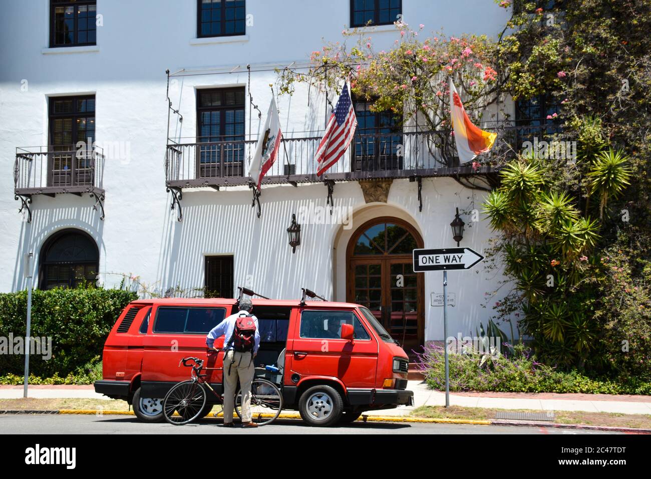 Rückansicht eines älteren Mannes, der das Fahrrad in den alten roten Volkswagen Vanagon lad, vor einem schönen Gebäude im spanischen Stil in Santa Barbara, CA, USA Stockfoto