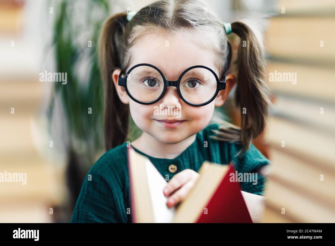 Nettes lächelndes Mädchen im Vorschulalter mit Grübchen in runden großen Gläsern mit rotem Buch in ihren Händen, das auf eine Kamera schaut Stockfoto