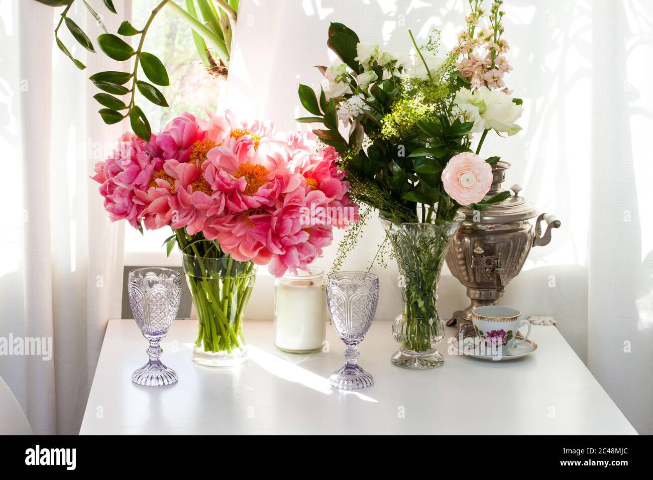 Blumenstrauß mit Pfingstrosen und Rosen, schwarze und weiße Kerzen, Weingläser und alte Teekanne auf weißem Tisch am Fenster. Stockfoto