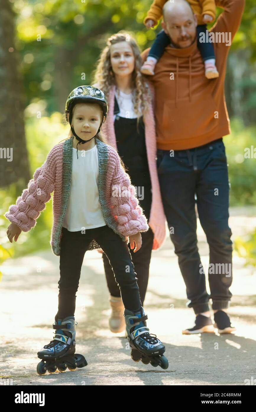 Junge glückliche Familie von Eltern und zwei kleine Kinder zu Fuß im Wald - ein Mädchen auf den Rollen Schlittschuh. Mittlerer Schuss Stockfoto