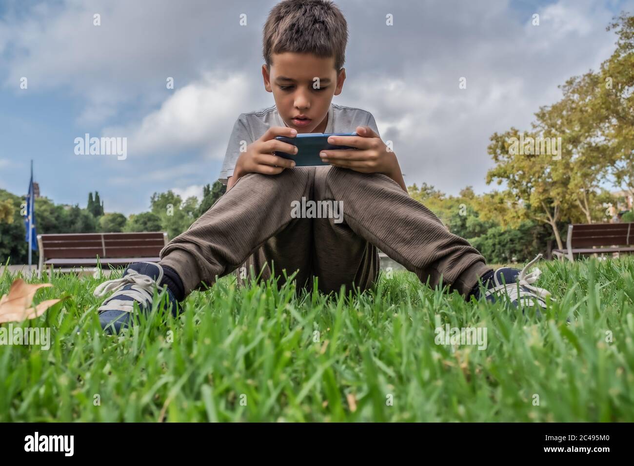Konzentrierter braunhaariger Junge, der im Park sitzt und mit einem Smartphone Spiele spielt. Stockfoto