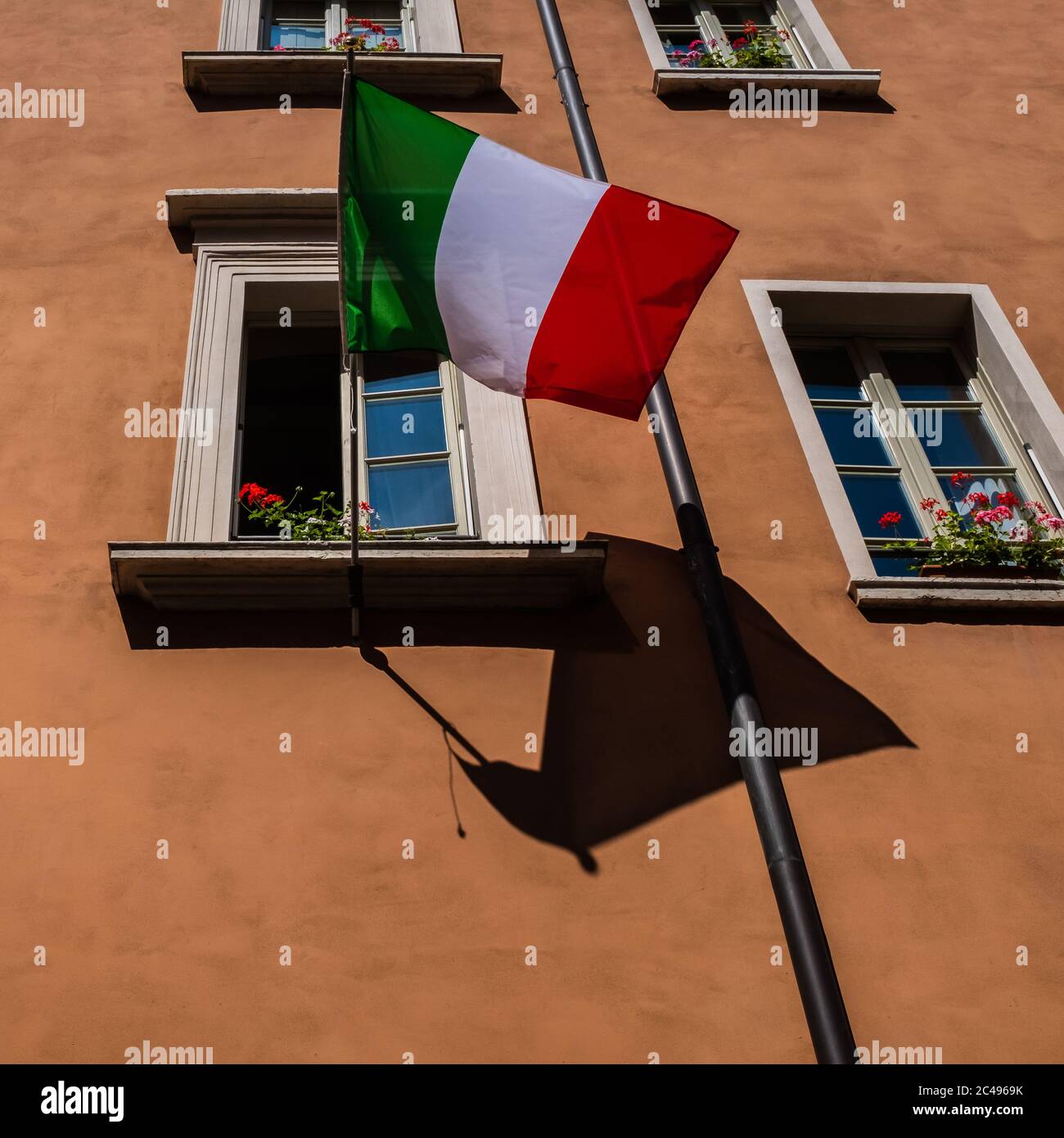 Nahaufnahme einer italienischen Flagge auf einer Fensterbank mit roten und weißen Blumen.die Fassade des alten Gebäudes ist ockerfarben gestrichen. Stockfoto