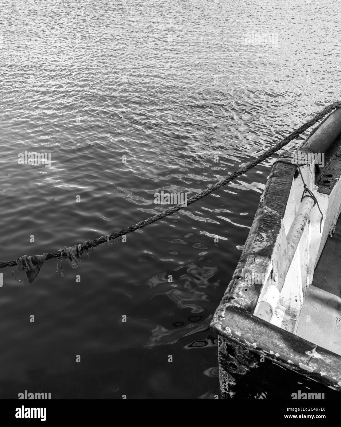 Schwarz-weißes Bild an einem Kai, wo Anlegeseile mit dem Heck eines rostenden Bootes verbinden. Die Lackierung ist abgesplittert und verkratzt. Stockfoto