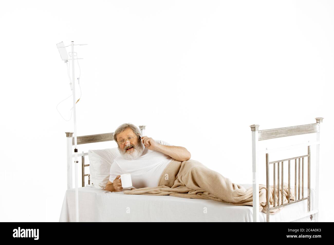 Älterer alter Mann erholt sich in einem bequemen Krankenhausbett isoliert auf weißem Hintergrund. Behandlung erhalten. Konzept der Gesundheitsversorgung und Medizin. Liegen, Telefonieren, Tee trinken. Copyspace. Stockfoto
