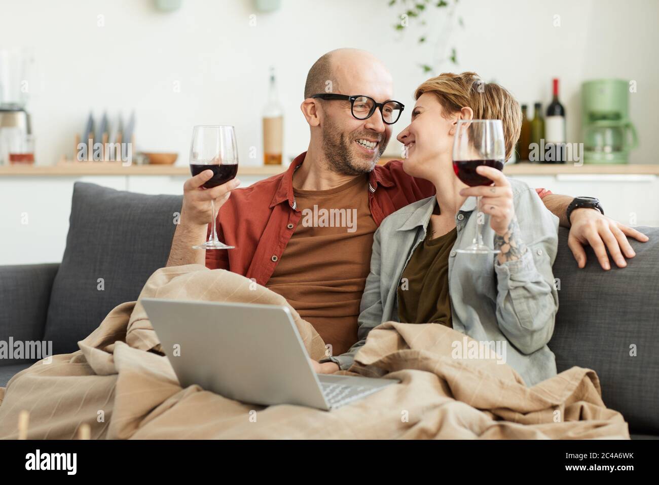 Glückliches reifes Paar, das auf dem Sofa mit Gläsern Rotwein am Laptop sitzt und lacht Stockfoto