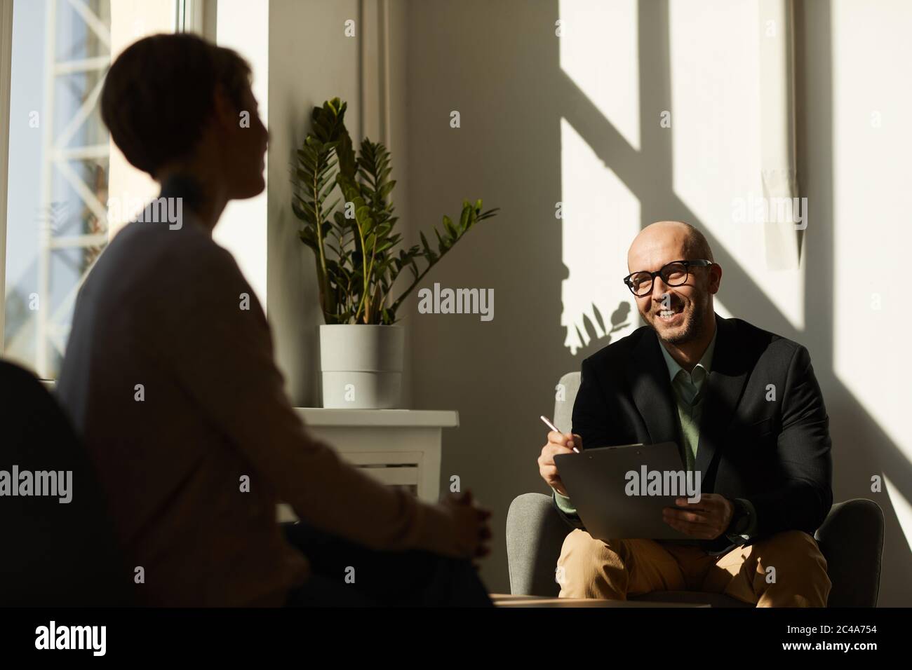 Reifer Mann in Brillen sitzt auf dem Sofa mit Laptop und lächelt, während sie mit Frau im Büro sprechen Stockfoto