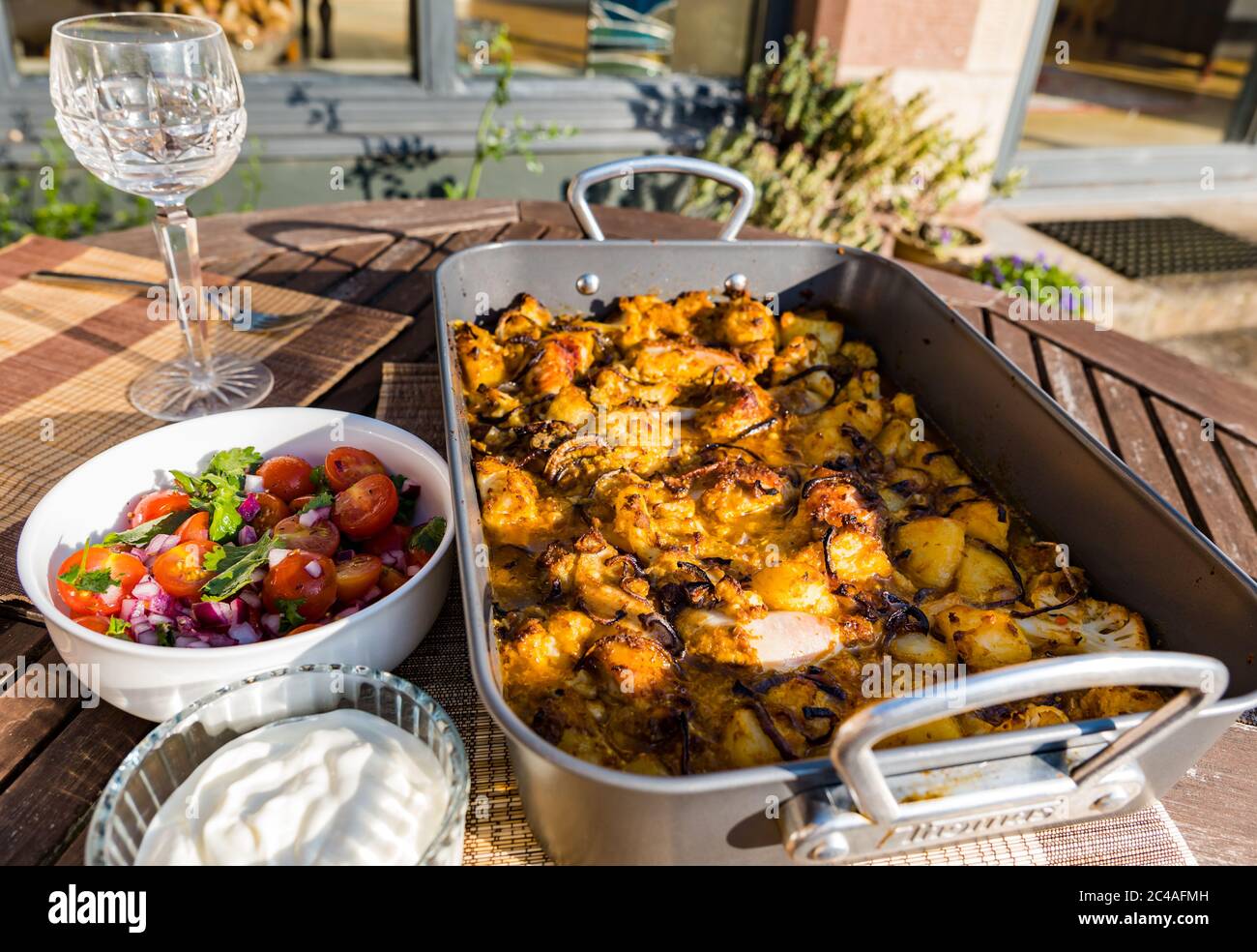 Chicken Aloo Gobi Curry serviert in Bräter Blech Backen Sie für Abendessen im Freien mit Tomaten und Minze Chutney und Joghurt in Sunshine, Großbritannien Stockfoto