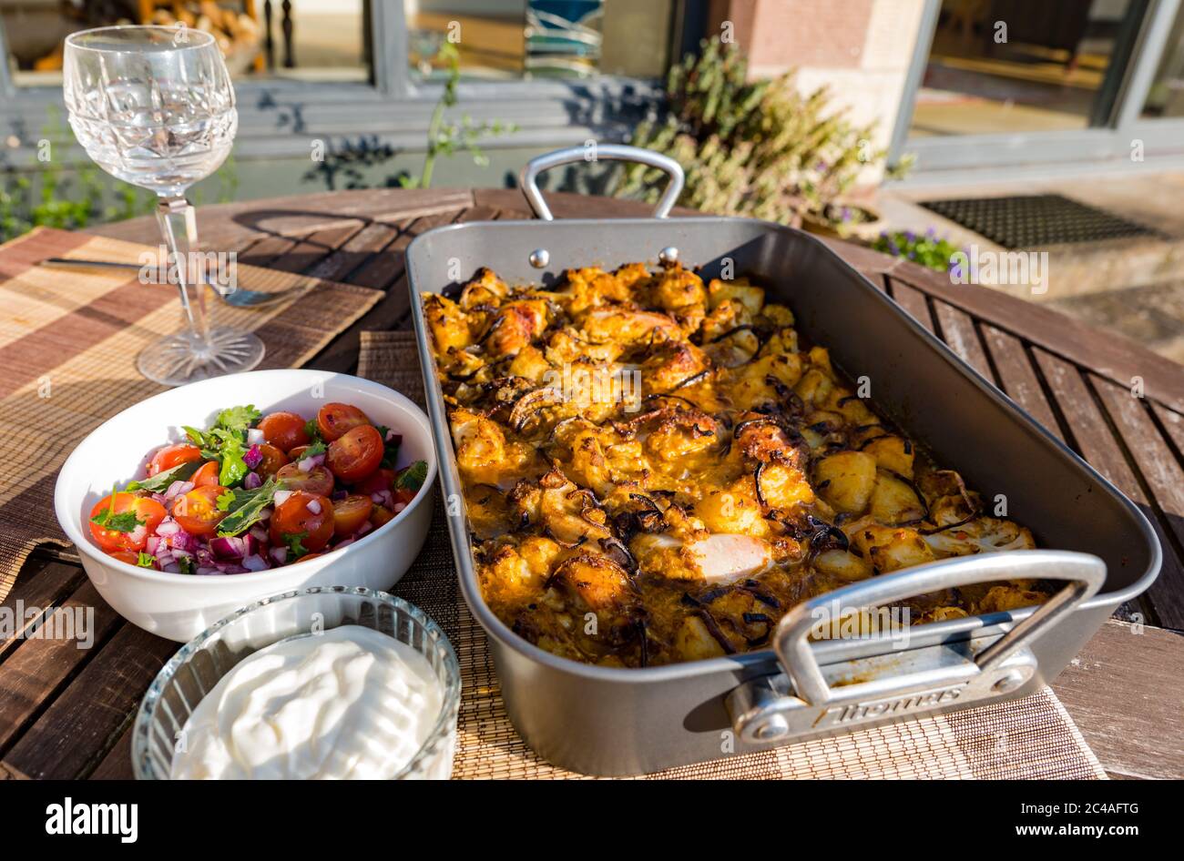 Chicken Aloo Gobi Curry serviert in Bräter Blech Backen Sie für Abendessen im Freien mit Tomaten und Minze Chutney und Joghurt in Sunshine, Großbritannien Stockfoto