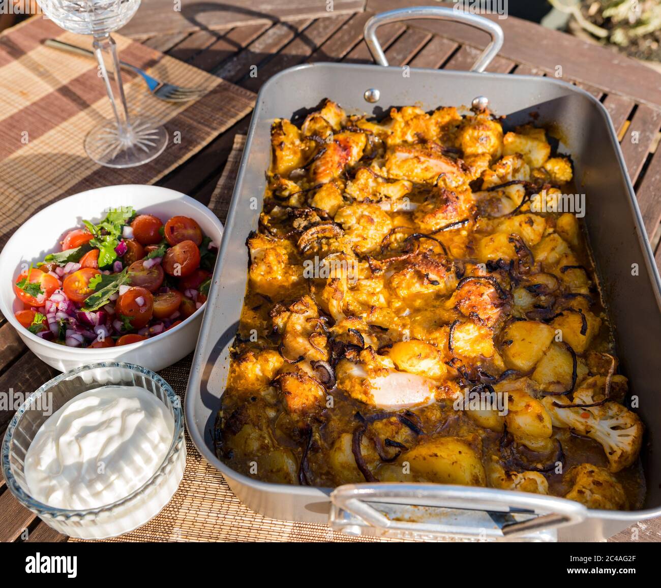 Chicken Aloo Gobi Curry serviert in Bräter Blech Backen Sie für Abendessen im Freien mit Tomaten und Minze Chutney und Joghurt in Sunshine, Großbritannien Stockfoto