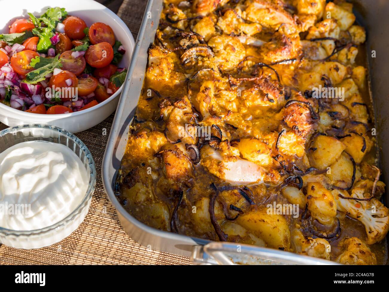 Chicken Aloo Gobi Curry serviert in Bräter Blech Backen Sie für Abendessen im Freien mit Tomaten und Minze Chutney und Joghurt in Sunshine, Großbritannien Stockfoto