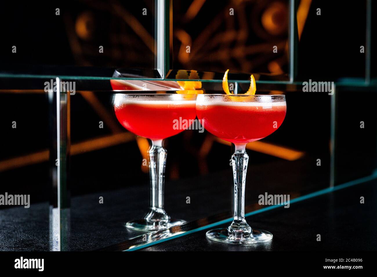 Ein rot-saurer Cocktail in einem Coupé-Glas, garniert mit einer Orangenschale. Spiegelung in einer Glaswand. Stockfoto