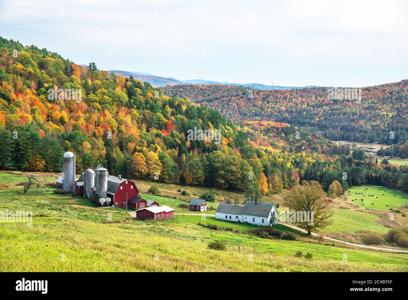 Hügelige ländliche Landschaft mit einem Bauernhof am Fuße eines Gebirges, bedeckt von Laubbäumen auf dem Gipfel des Herbstlaubes an einem bewölkten Herbsttag Stockfoto