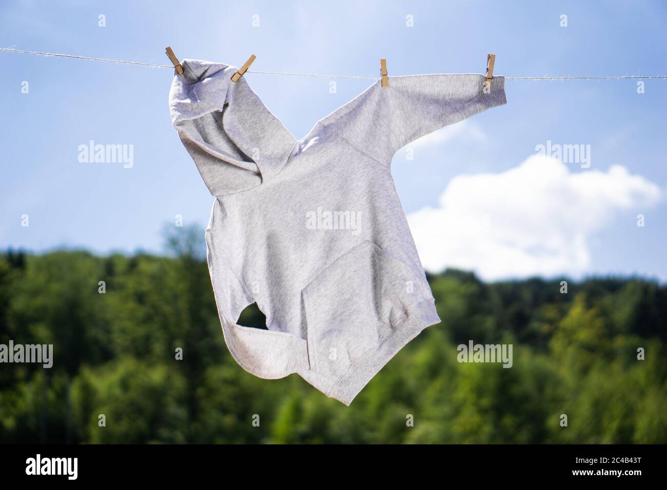 Graues Kapuzenpullover, das am Seil mit Wäscheklammern vor blauem Himmel und grünen Bäumen im Sommer-Hintergrund hängt. Modellkonzept. Stockfoto
