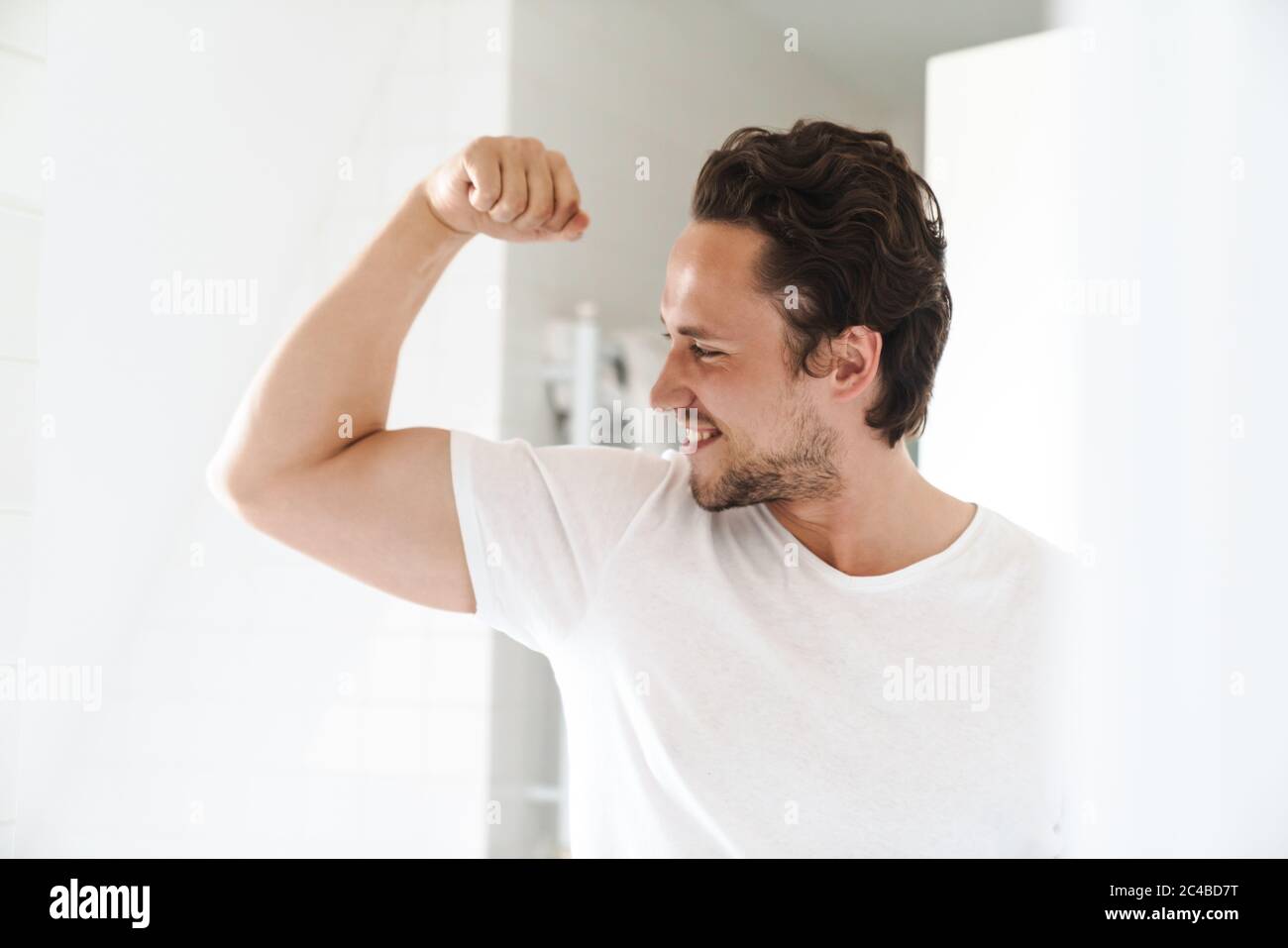 Attraktiver, glücklicher, selbstbewusster junger Mann, der vor dem Badezimmerspiegel steht und Bizeps beugt Stockfoto
